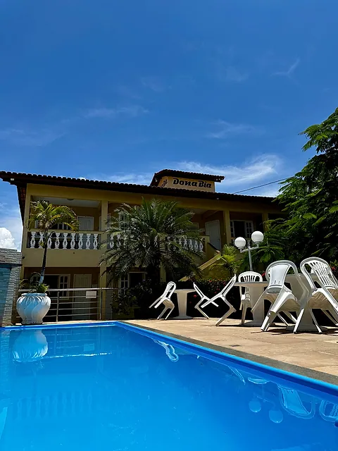 Piscina com cadeiras brancas e o prédio da pousada ao fundo